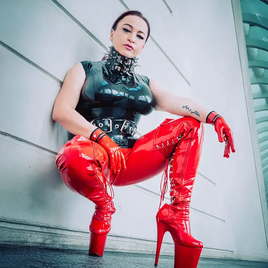Mistress Oksana in dominanter Hocke im Golden Cage Dominastudio Aachen. Sie trägt ein schwarzes Latex-Top mit Stachelhalsband, rote Latex-Leggings und rote Plateau-Overknee-Stiefel im professionellen Domina Studio.